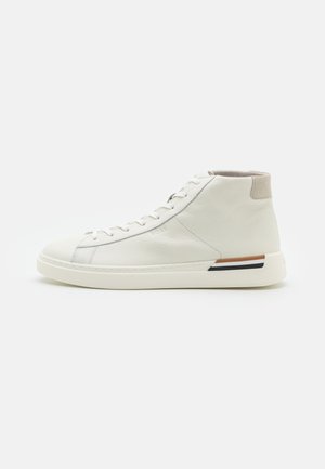 Zapatilla deportiva alta blanca de cuero con cordones, collar acolchado beige en el tobillo y suela blanca con franjas negras y marrones en el talón.