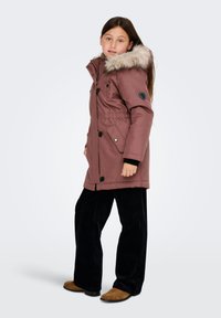 Rózsaszín parka, műszőrme kapucnival, elasztikus derékkal, fekete kiegészítőkkel és patent/zárós gombbal. A modellen fekete kordbársony nadrág és barna cipő van.