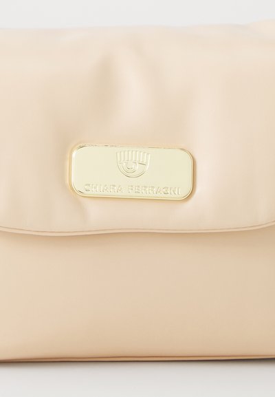 Borsa a tracolla in pelle beige con pattina pieghevole e una placca logo in metallo dorato con testo. Texture liscia con dettagli minimi.