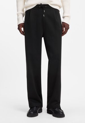 Uomo che indossa pantaloni neri a gamba larga con coulisse in vita, scarpe nere e maglione bianco a coste con maniche lunghe.