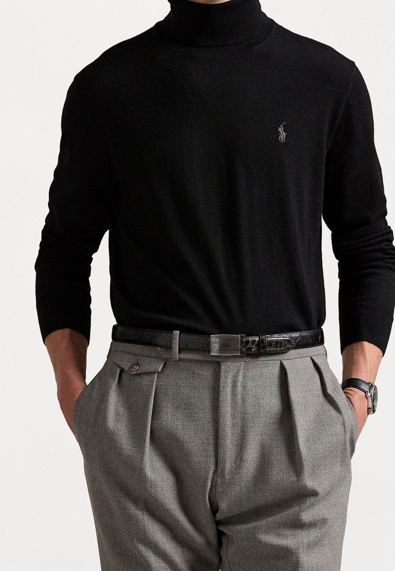 Pull à col roulé noir avec des manches longues, associé à un pantalon gris plissé et une ceinture noire à motifs. Montre visible au poignet.