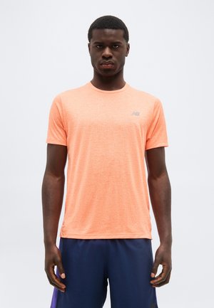 Mladý muž stojí čelom k fotoaparátu, má na sebe broskyňové športové tričko s krátkym rukávom a tmavomodré šortky, na jednoduchom svetlom pozadí.