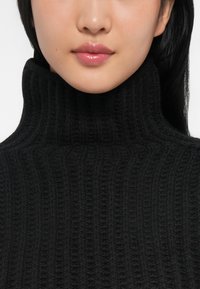 Mlada žena s glatkom kožom i sjajnim usnama nosi crni rebrasti džemper s turtleneck izrezom, otkrivajući vrat i donji dio lica.