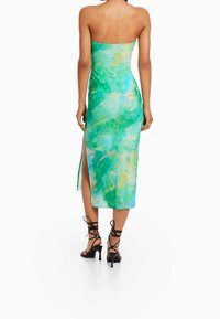 Femme portant une robe midi sans bretelles en tie-dye vert et bleu avec une fente latérale et des sandales à talons hauts noires à lacets, se tenant de dos.