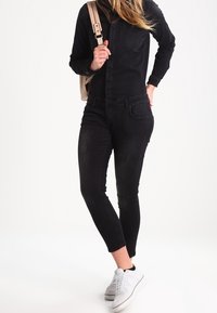 Černá denimová košile a skinny džíny v kombinaci s bílými teniskami; má přední kapsu a světlý batoh.