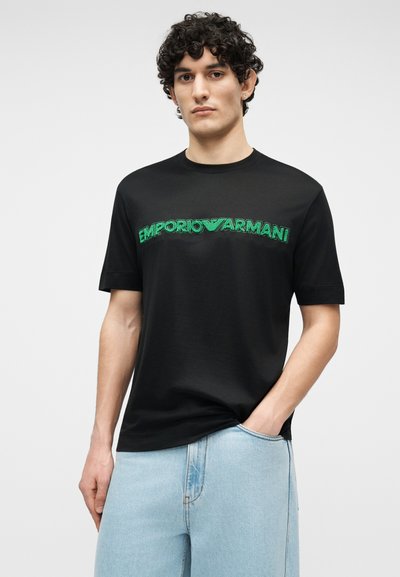 Νεαρός άνδρας με σγουρά μαλλιά, που φοράει μαύρο T-shirt με πράσινο λογότυπο "Emporio Armani" και ανοιχτό μπλε τζιν, με το ένα χέρι στην τσέπη.