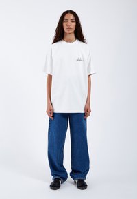 Camiseta de algodón blanca con cuello redondo y mangas cortas, que presenta un logo negro escrito a mano. Combinada con jeans de denim azul sueltos y zapatos negros.