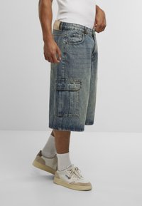 Denim kratke hlače, modre z zbledelim učinkom, z velikimi stranskimi žepi in ravnim krojem, v kombinaciji z belimi športnimi copati in nogavicami do gležnjev.