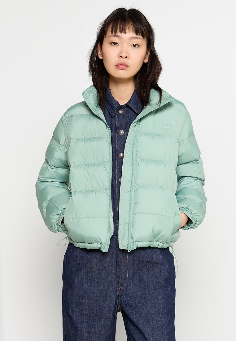 Chaqueta acolchada verde claro con cierre de cremallera en la parte delantera, botones a presión, puños elásticos y un cuello alto. Acabado texturizado con relleno acolchado.