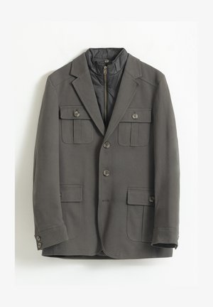 Blazer gris con una capa interna acolchada y con cremallera. Presenta dos bolsillos en el pecho, dos bolsillos frontales y cierres de botón. Textura suave.