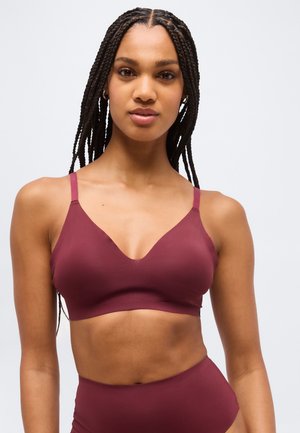 Ženska z pletenicami, oblečena v brezšivni bordo barve nedrček in ujemajoče se hlačke z visokim pasom, na svetlem ozadju.