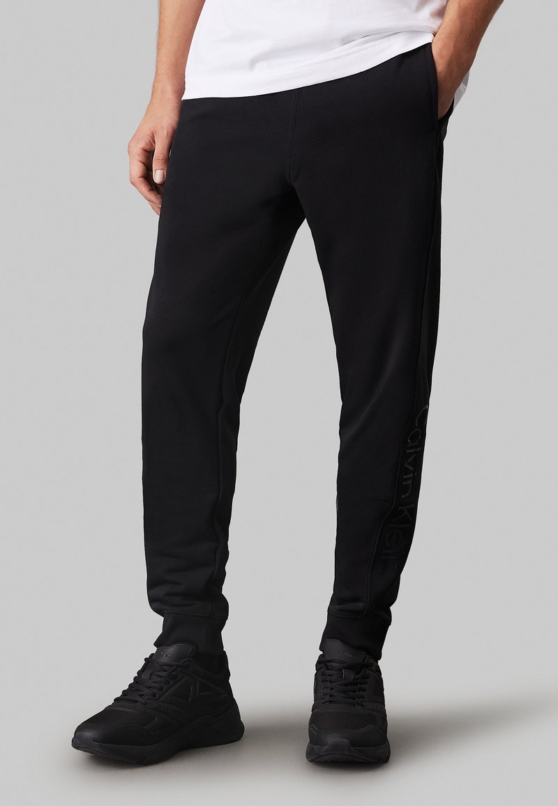 Pantaloni da jogging neri con una vestibilità slim, realizzati in materiale morbido. Presentano una stampa del logo laterale e polsini a costine, abbinati a scarpe sportive nere.