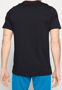T-shirt en coton noir à manches courtes, col rond et ourlet droit ; porté avec un short de sport bleu montrant des détails latéraux contrastants.