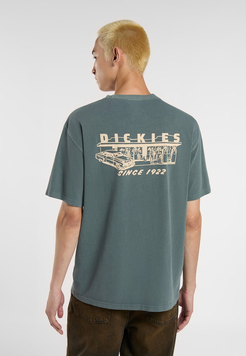 Camiseta de manga corta en color teal con un diseño gráfico beige de una gasolinera vintage y un coche en la parte posterior, con el texto "DICKIES" arriba.