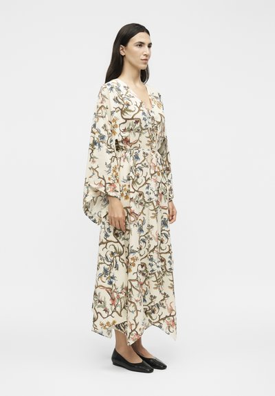 Γυναίκα στέκεται φορώντας ένα μακρύ κρεμ  φόρεμα με λουλουδάτο σχέδιο, φαρδιά μανίκια και μαύρα επίπεδα παπούτσια, στραμμένη προς τα δεξιά σε ένα απλό λευκό φόντο.