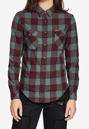 Persona con una camisa de cuadros abotonada de manga larga con cuadros burdeos y verdes y pantalones negros con agujeros remendados.