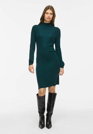 Femme portant une robe en maille ajustée vert foncé à manches longues avec un col montant et des bottes noires montant jusqu'aux genoux, debout devant un fond blanc uni.