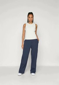 Nainen seisoo kädet taskuissa, päällään valkoinen ilman hihoja oleva toppi, väljät tummansiniset housut ja valkoiset tennarit, yksinkertaista vaaleaa taustaa vasten.