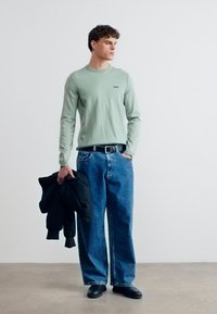 Άνδρας που φοράει ανοιχτό πράσινο πουλόβερ και φαρδιά μπλε τζιν, κρατώντας μια μαύρη ζακέτα, στέκεται μπροστά σε έναν απλό λευκό τοίχο.