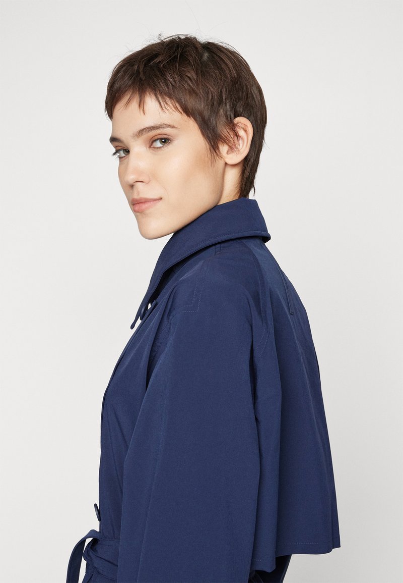 Jeune personne avec les cheveux courts et bruns portant une veste à col bleu foncé, regardant par-dessus son épaule contre un fond clair. 