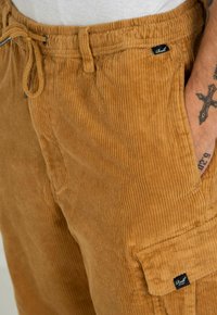 Brown corduroy pants feature a drawstring waist, side pocket, and a small black label on the front, showcasing vertical ribbed texture.