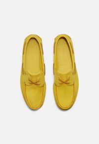 Ett par gula lädermoccasin-loafers med bruna snördetaljer, sedda ovanifrån på en vit bakgrund.