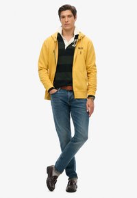 Sudadera amarilla con cremallera, polo a rayas negro y verde, pantalones vaqueros azules, cinturón de cuero marrón, zapatos oscuros, pose de pie con las manos en los bolsillos.