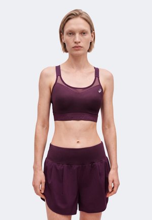 Frau trägt einen dunkelvioletten Sport-BH und passende Sportshorts, steht mit entspannten Armen vor einem schlichten hellen Hintergrund.