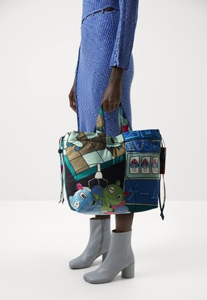 Persona con un vestido de punto azul de canalé y botines de tacón gris claro sosteniendo un bolso tote colorido con personajes de dibujos animados y motivos de juegos.