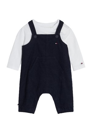 Salopette en velours côtelé bleu marine avec poche avant, portée sur une chemise blanche à manches longues, conçue pour les nourrissons ou tout-petits avec bretelles et détails à boutons.