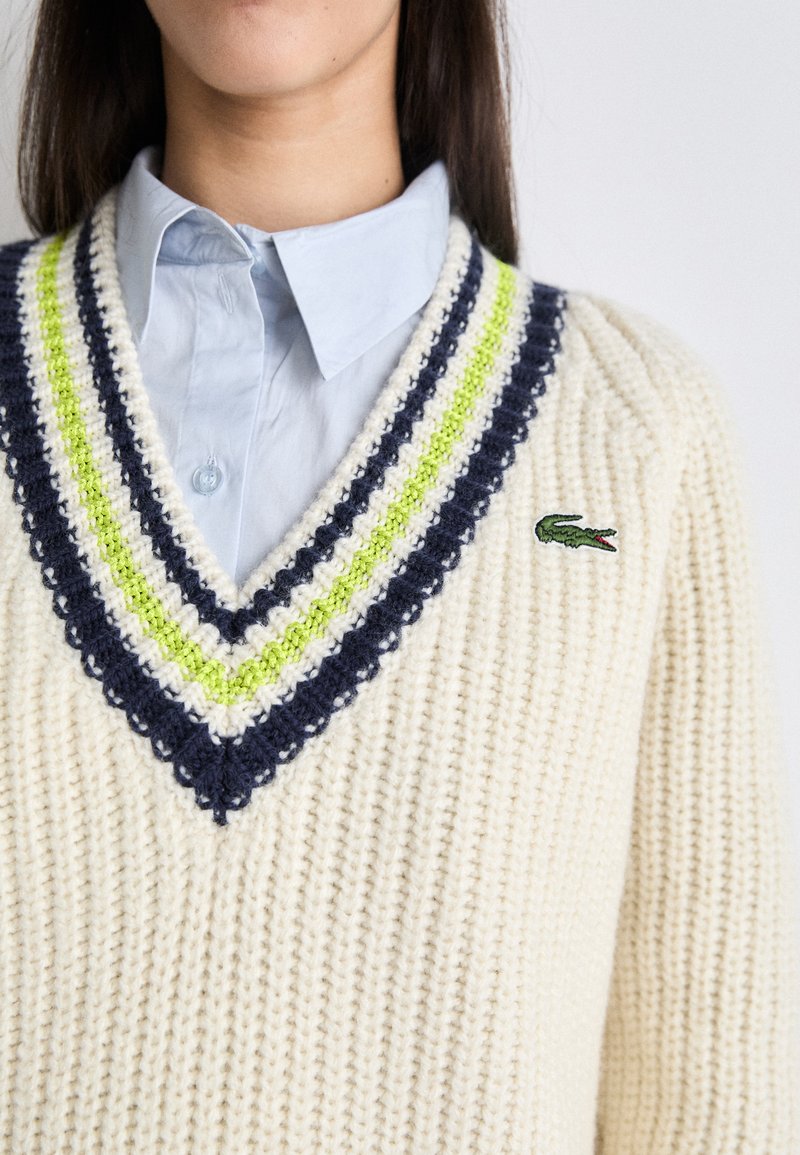 Pull en tricot couleur crème avec un col en V, doté d'accents rayés bleu marine et vert lime, et d'un logo crocodile vert sur la poitrine.