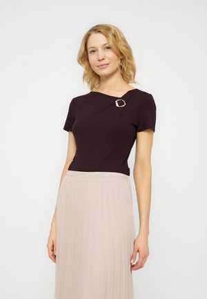 Femme aux cheveux blonds portant un haut noir à manches courtes avec une broche et une jupe plissée beige clair, debout devant un fond uni.