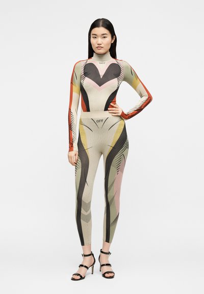 Γυναίκα που φοράει εφαρμοστό ολόσωμο με πολύχρωμο γεωμετρικό σχέδιο και μακριά μανίκια, με μαύρα πέδιλα με τακούνια, στέκεται με το ένα χέρι στη μέση.