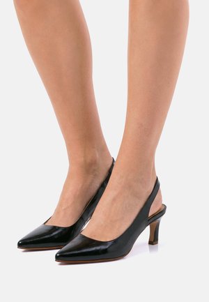 Pies de mujer con slingback de cuero negro y tacones kitten puntiagudos sobre un fondo blanco.