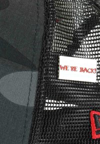 Black cap featuring a mesh back. The front displays a rectangular pin with the text "WE'RE BACK!" in red. Textured fabric and pattern visible.