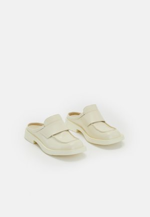 Beige Slipper mit einer runden Zehenpartie, einem breiten Riemen über dem Vorderfuß und einem klobigen Absatz. Das Material wirkt glatt und glänzend.