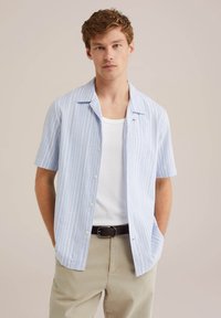 Chemise à manches courtes avec boutonnière en bleu clair à rayures verticales blanches. Présente un col classique et une coupe décontractée. Associée à un sous-vêtement blanc.