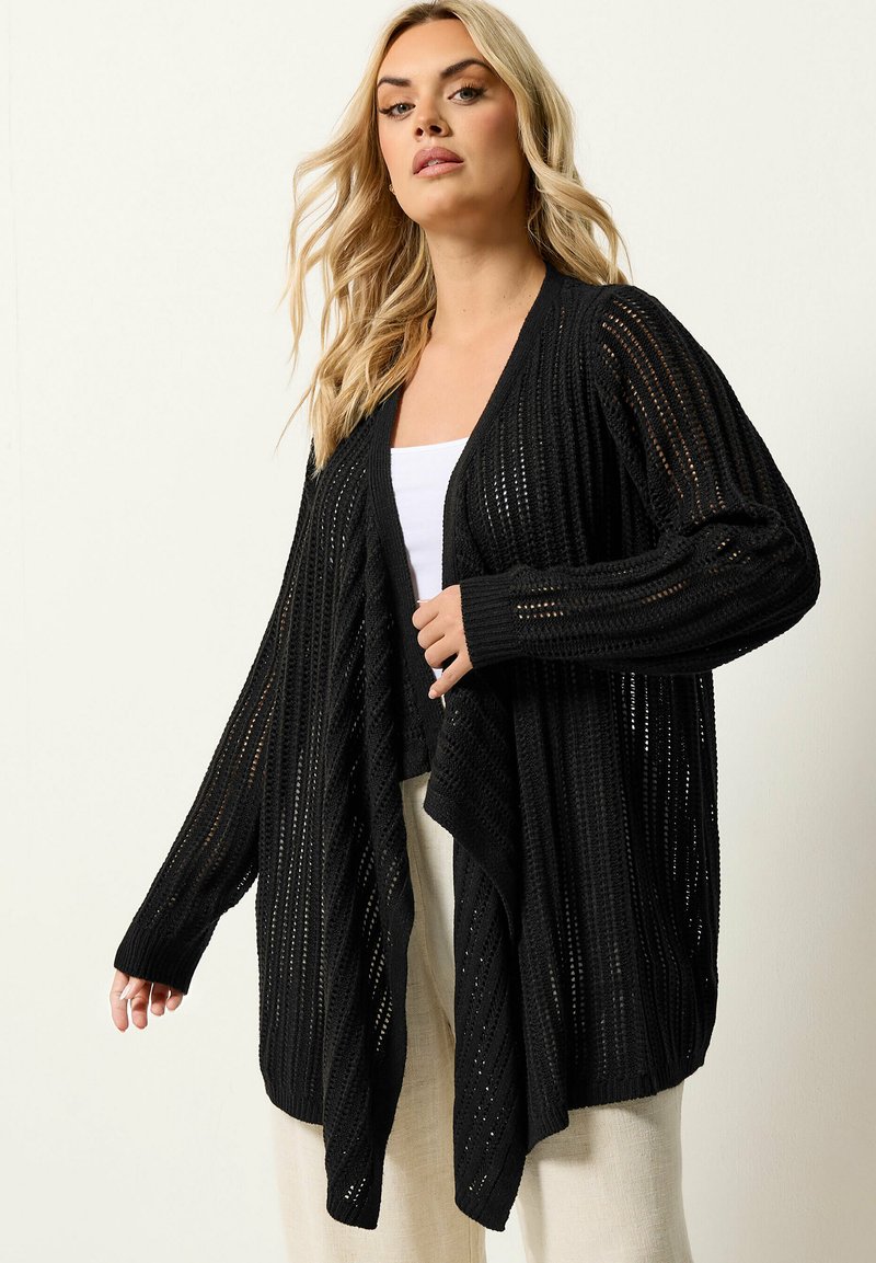 Blonde Frau, die einen schwarzen, offen gestrickten Cardigan über einem weißen Top und hellbeigen Hosen trägt, vor einem schlichten Hintergrund stehend.