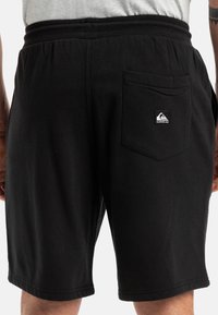 Pantalones cortos negros de tejido suave, con cintura ajustable mediante cordón, bolsillos laterales y un bolsillo trasero con una pequeña etiqueta de logo.