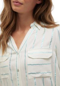 Chemise blanche à boutons avec des rayures bleues, dotée de deux poches sur la poitrine et d'un col. Tissu léger, texture lisse, design simple.