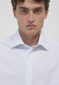 Jeune homme aux cheveux bouclés portant une chemise blanche à fines rayures verticales bleues et au col ouvert, faisant face à l'avant.