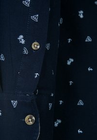 Marinblus med vita nautiska mönster, med knappar och en slät textur. Närbild på manschett och sid sömnadsdetaljer.