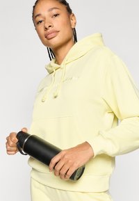 Κίτρινο φούτερ με κουκούλα, κορδόνια και μπροστινή τσέπη, κρατιέται με μαύρο μονωμένο μπουκάλι νερού. Διαθέτει λεία υφή και ελάχιστη σήμανση.