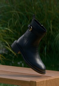 Černá kotníková bota z texturované ků�že se zlatou sponou, vznášející se nad dřevěným povrchem, v pozadí rozmazaná zeleň.