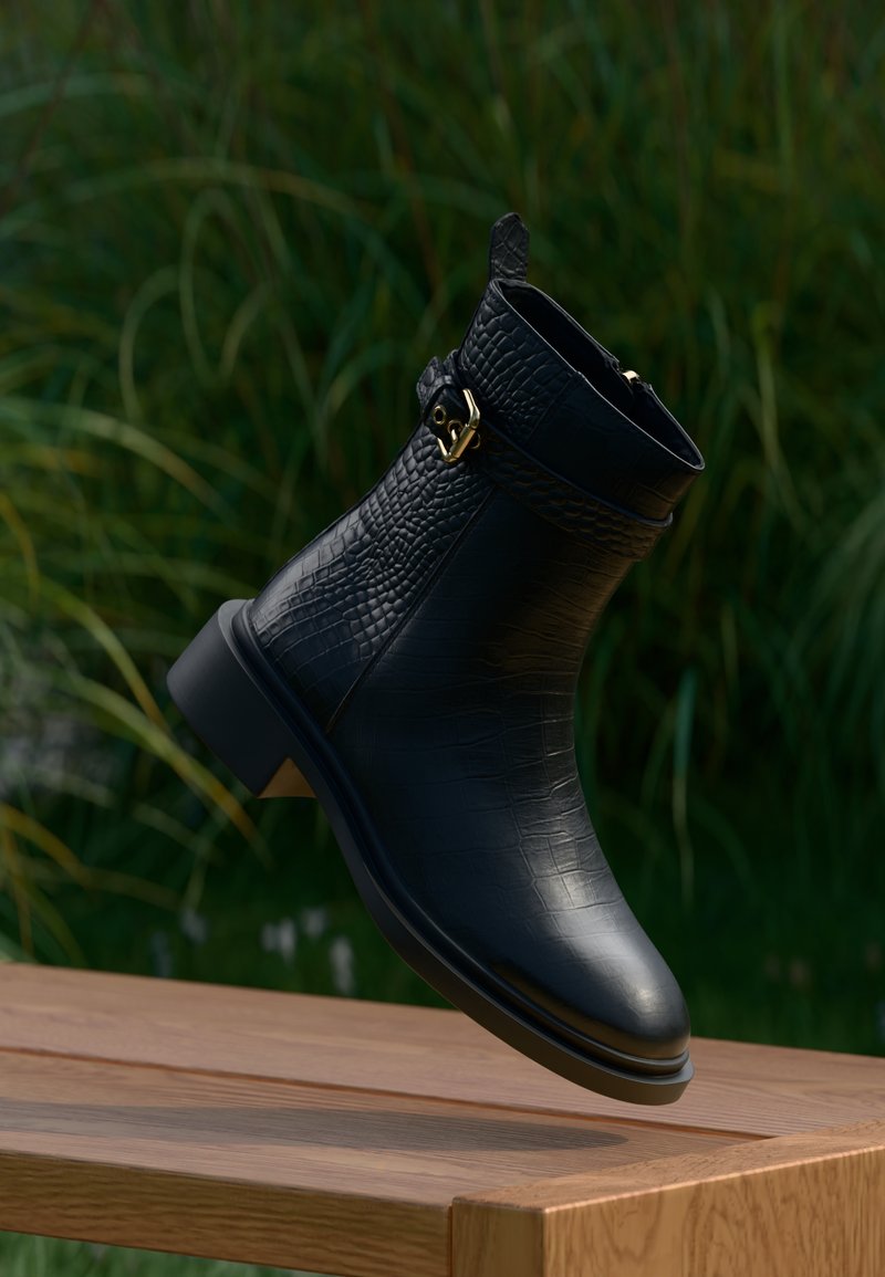 Bota preta de tornozelo em pele texturada com fivela dourada a pairar sobre uma superfície de madeira, com folhagem desfocada ao fundo.