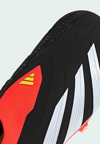 Chaussure de soccer noire avec accents rouges, dotée de trois rayures blanches, d'une tige texturée et d'un détail de logo jaune sur le côté près du talon.