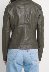 Kurtka skórzana w oliwkowej zieleni o dopasowanym kroju, z widocznymi szwami i gładką teksturą. Zestawiona z jasnoniebieskimi jeansami.