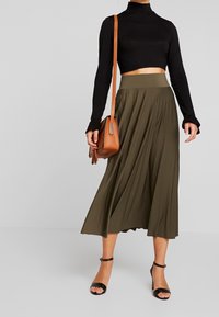 Gonna midi plissettata verde oliva con vita alta, abbinata a un top nero aderente a maniche lunghe e sandali neri. Borsa a tracolla marrone inclusa.