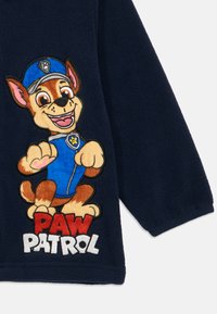 Μανίκι από μπλε φλις με κεντημένο καρτούν σκύλο σε μπλε στολή και μπλε καπέλο πάνω από το κόκκινο και λευκό κείμενο "PAW PATROL".