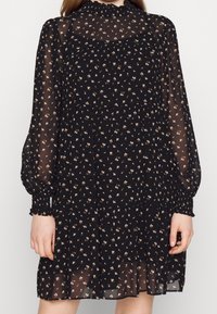 Schwarzes Blumenkleid aus transparenter Stoff, mit langen Puffärmeln und einem rüschierten Kragen. Kleine weiße und beige Blumen bilden ein wiederholendes Muster.